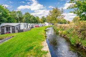 Riverside touring pitches with caravans and awnings facing a stream and dry-stone wall.