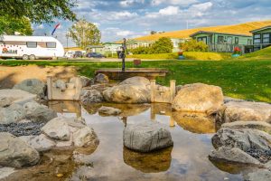 Shallow stream with stepping stones running through our Lake District caravan park, with green fells and holiday homes beyond.